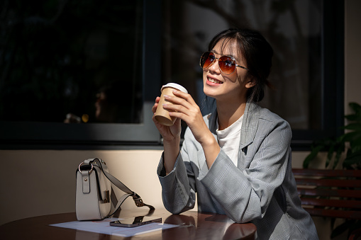 Young beautiful and happy Asian woman wearing sunglasses enjoying her coffee at a cafe on a sunny day. city life concept