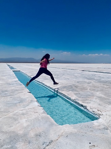 A young woman caught in-motion leaping over pristine blue water in the salt flats of Salta in Argentina, called Salinas Grandes.