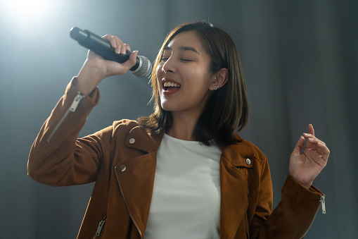 Attractive singer woman singing music on stage with spotlight strike through the darkness at concert event. Musician performing live with light in hall.