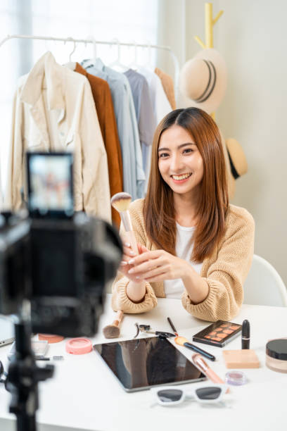 istock Young beautiful asian woman vlogger live broadcasting cosmetic makeup tutorial content with brush on looking at cell phone. Influencers with various cosmetic recording review video sharing on social 1941435543