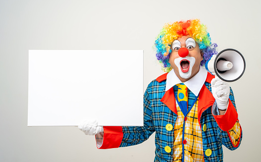 Funny clown in a multi colored wig with his thumbs up leaning over a white blank sign. He is wearing golden dollar shaped sunglasses. Isolated on white. You can add extra white space with your message to the bottom.