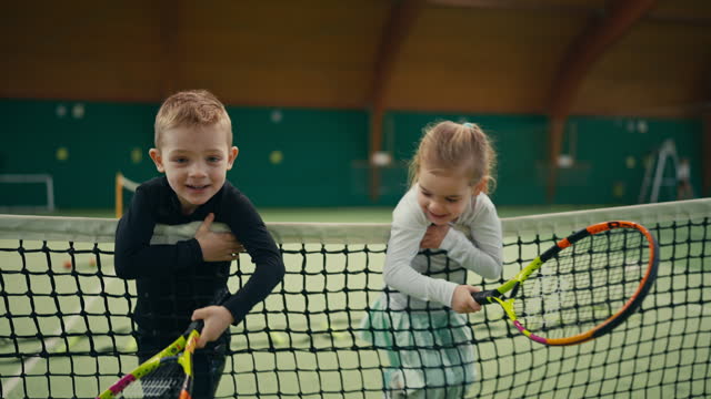 Portrait of Playful Little Children with Tennis Rackets Running and Leaning on Net in Sports Club