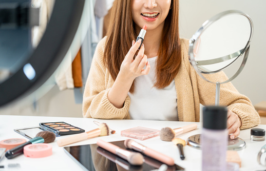 Young beautiful asian woman vlogger live broadcasting cosmetic makeup tutorial content with lipstick looking at cell phone. Influencers with various cosmetic recording review video sharing on social
