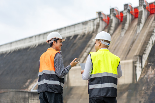 Confident asian two maintenance engineers man inspection discussstion with tablet at construction site dam with hydroelectric power plant and irrigation. Team engineer man working at project