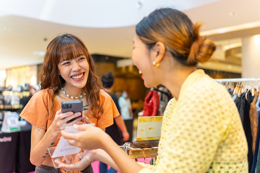 Young Asian woman buying handmade jewelry and using smartphone scan QR code for making online at shopping mall. Small business shop owner and contactless payment technology concept.
