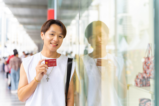 Portrait of Young Asian man holding credit card standing in front of shopping mall in the city. Generation z people enjoy shopping with contactless payment.