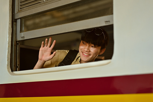 Cheerful young Asian man waving hand to his friend through train window. Travel and lifestyle concept.