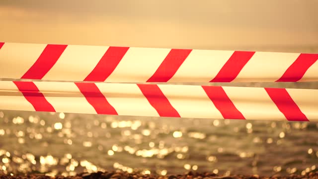 Red white warning tape barrier ribbon swinging in the wind across exotic sea beach background without people. No entry Red White caution tape. No holiday concept, delayed travel, no summer plans