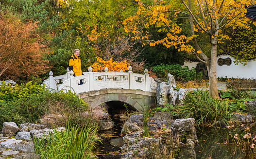 Fall scene in Midwestern park with senior man standing on stone bridge over landscaped stream; trees in background