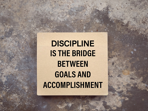 DISCIPLINE IS THE BRIDGE BETWEEN GOALS AND ACCOMPLISHMENT written on a notepad. With blurred style background.