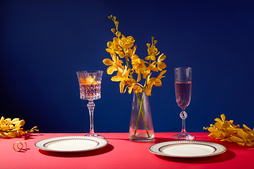 Yellow orchid branches are placed in a glass vase, two wine glasses contain light pink liquid, and there are two porcelain plates on the table. Mysterious dark blue background.