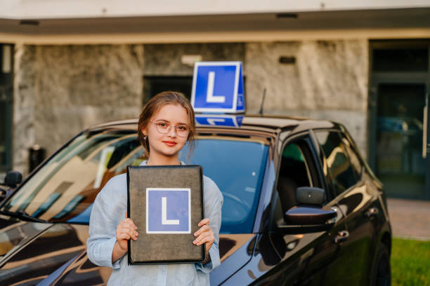 giovane autista femminile affascinante in posa con la piastra l sul tetto del veicolo. scuola guida e concetto di persone. giovane bella donna felice che tiene la cartella con il segno l vicino all'auto di addestramento. - driving training car safety foto e immagini stock