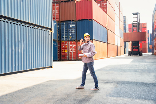 young asian man walking and holding walkie talkie to communicate while working in cargo distribution at commercial dock