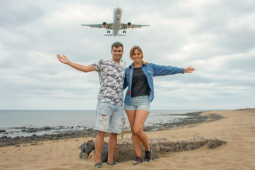 Couple on a beach with open arms and a commercial airplane on top of them.