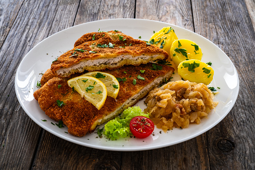 Breaded fried pork chop with boiled potatoes and fried cabbage on wooden background