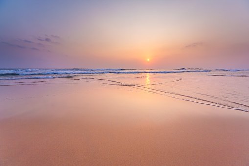 A serene scene with the sun descending over the ocean, casting warm hues on wet sand and gentle waves. The calm waters reflect the sky’s colors, creating a peaceful ambiance at this secluded beach during sunset. Shot taken in hikkaduwa.
