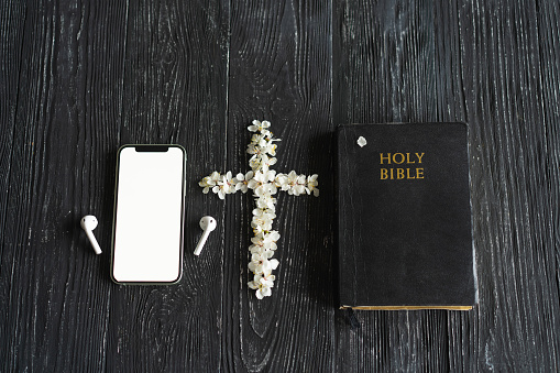 Spring flowers in the shape of a cross, a phone with headphones and holy bible on an old wooden background.
