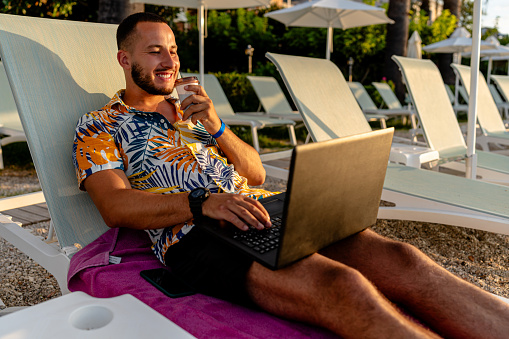 A picturesque beach setting, the aroma of his coffee, and pressing tasks on his laptop - a young man perfectly encapsulates the balance of vacation and vocation