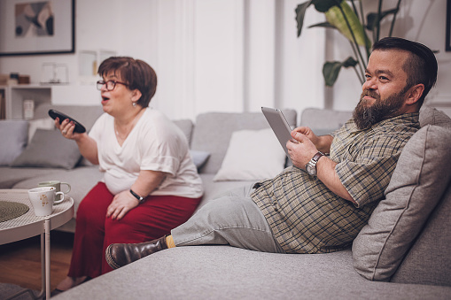 A woman with a TV remote and a man with a tablet share a relaxing time in the living room, engaged in leisure activities