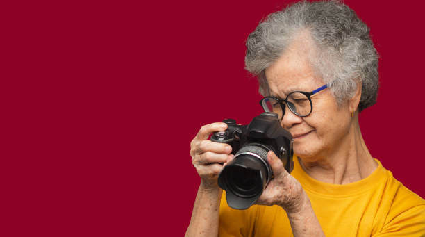 Happy senior woman taking photographs with a digital camera while standing on a red background. Aged people photographer and relaxation concept