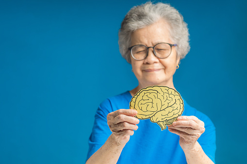 Hands of a senior woman holding a brain shape made from yellow paper against a blue background. Alzheimer's, Parkinson's disease, dementia, stroke, seizure, or mental health. Healthcare concept