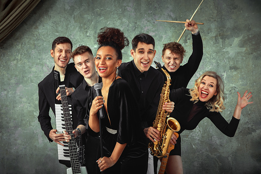 Cheerful international music group on a gray wall background, a group of musicians posing on camera in the hands of various instruments, guitars, saxophone. Copy space