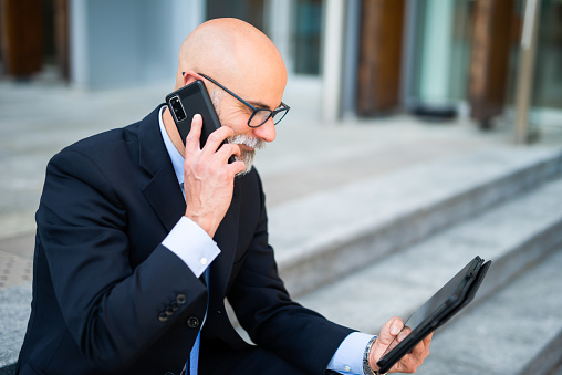 Bald businessman with white beard using his tablet while talking on the phone in a modern city