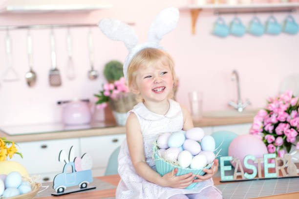 istock Cute little girl with bunny ears holding Easter egg in the kitchen decorated for Easter. 1942097778