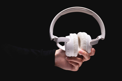 A woman hand holds large white headphones on a black background. A girl holds out wireless headphones to the camera, close-up