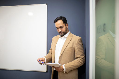 A businessman working by a whiteboard in a modern office. He is wearing a beige coloured jacket and white shirt.