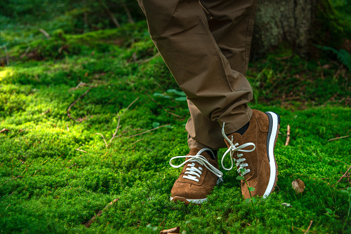 Male`s legs crossed at the ankle standing on moss wearing brown suede sneakers in the dense
forest