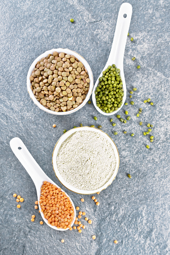 Lentil flour in bowl, red, green lentils in spoons, brown in bowl, bean stalks with leaves on stone table background from above