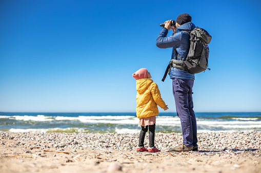 A tourist man standing on the seashore, using binoculars to observe the coastal landscape. His little daughter standing nearby.