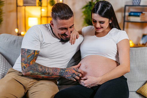 Beautiful young pregnant woman embracing with her husband while sitting on the sofa in the living room at night.