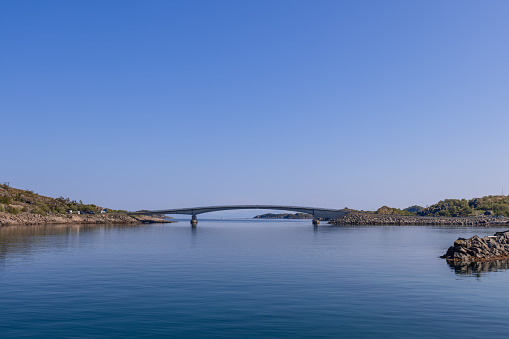 A slender bridge arches gracefully over the still waters of Lofoten, connecting rocky outcrops under a vast, clear blue sky in a serene Norwegian landscape