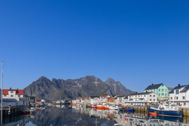 las aguas tranquilas reflejan el puerto de henningsvaer y heimsundet, flanqueadas por las escarpadas montañas de lofoten, noruega - lofoten henningsvaer norway village fotografías e imágenes de stock