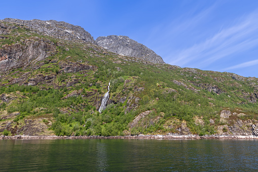 Lush greenery frames a slender waterfall in Norway's majestic Trollfjorden, under a streaked blue sky