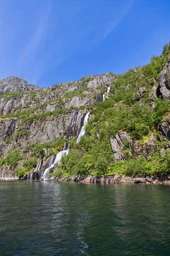 Fresh waterfalls cut through the lush, moss-covered cliffs of Trollfjorden, Lofoten, flowing into the tranquil waters below under a clear blue sky
