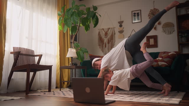 Mother using her laptop to watch online yoga classes and practice with her small daughter in the living room at home
