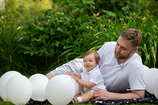 A dad and his little son lie on the lawn surrounded by white balloons. Father's Day.