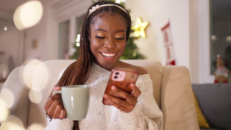 Woman Black ethnicity enjoying Christmas atmosphere, while drinking tea and using mobile phone