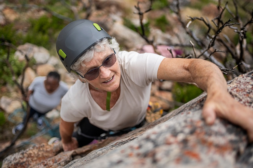 Top view of a healthy senior woman rock climbing on the cliff with female friend belaying standing in the bottom of the mountain