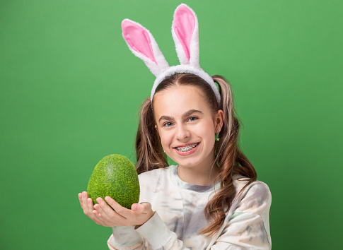 Happy Easter. Cheerful girl with Easter bunny ears and big Easter egg made of green grass on green background. Copy space. Smiling girl in braces.