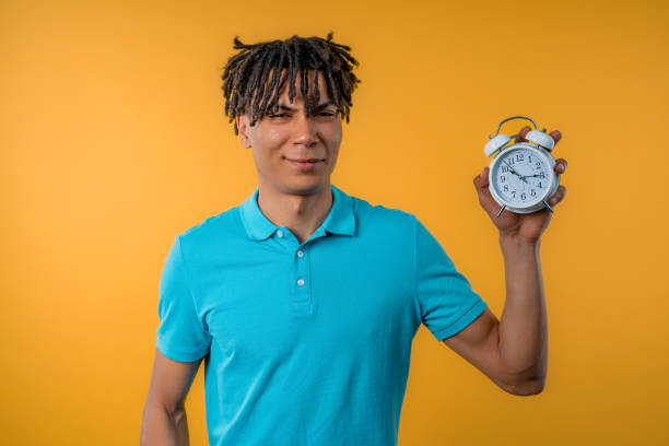 woken up by alarm clock sleepy african man holding it in hand. yellow background - clock time clock hand urgency photos et images de collection