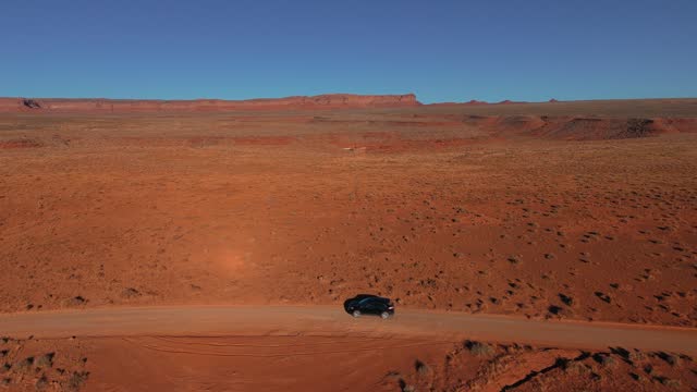 Car driving on a desert dirt road near Valley of the Gods and Monument Valley in Utah and Arizona. Modern black SUV automobile aerial drone point of view in cinematic red sandstone rock formation landscape. Blue sky in Western USA, Wild West movie scenery