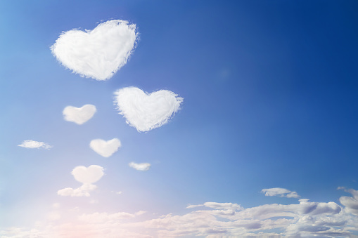 Fluffy clouds forming a hearts shape on blue sky with sun, soft focus. Heavenly clouds background. Valentine day, love, romantic, Mother day concept. Copy space. Empty place for message.
