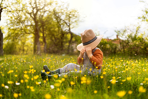 Little unrecognizable boy sitting in the spring grass and hiding his face under the sun hat.