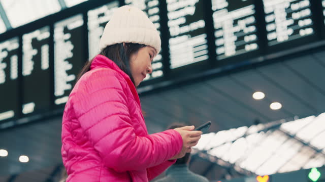 Asian woman passenger traveler looking at Arrival Departure Board status timetable and time schedule and using smart phone at train station public transportation
