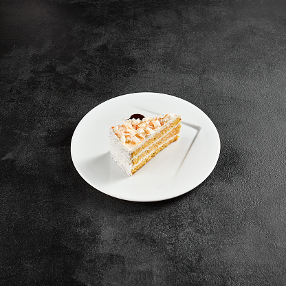 A slice of white chocolate and coconut cake on a white plate with dark syrup, against a textured black backdrop.