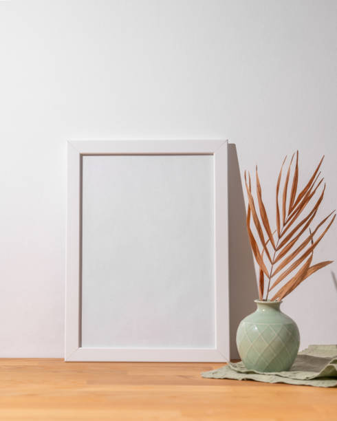 istock A white frame with a blank canvas against a light wall and on a wooden tabletop with vase. 1942608471
